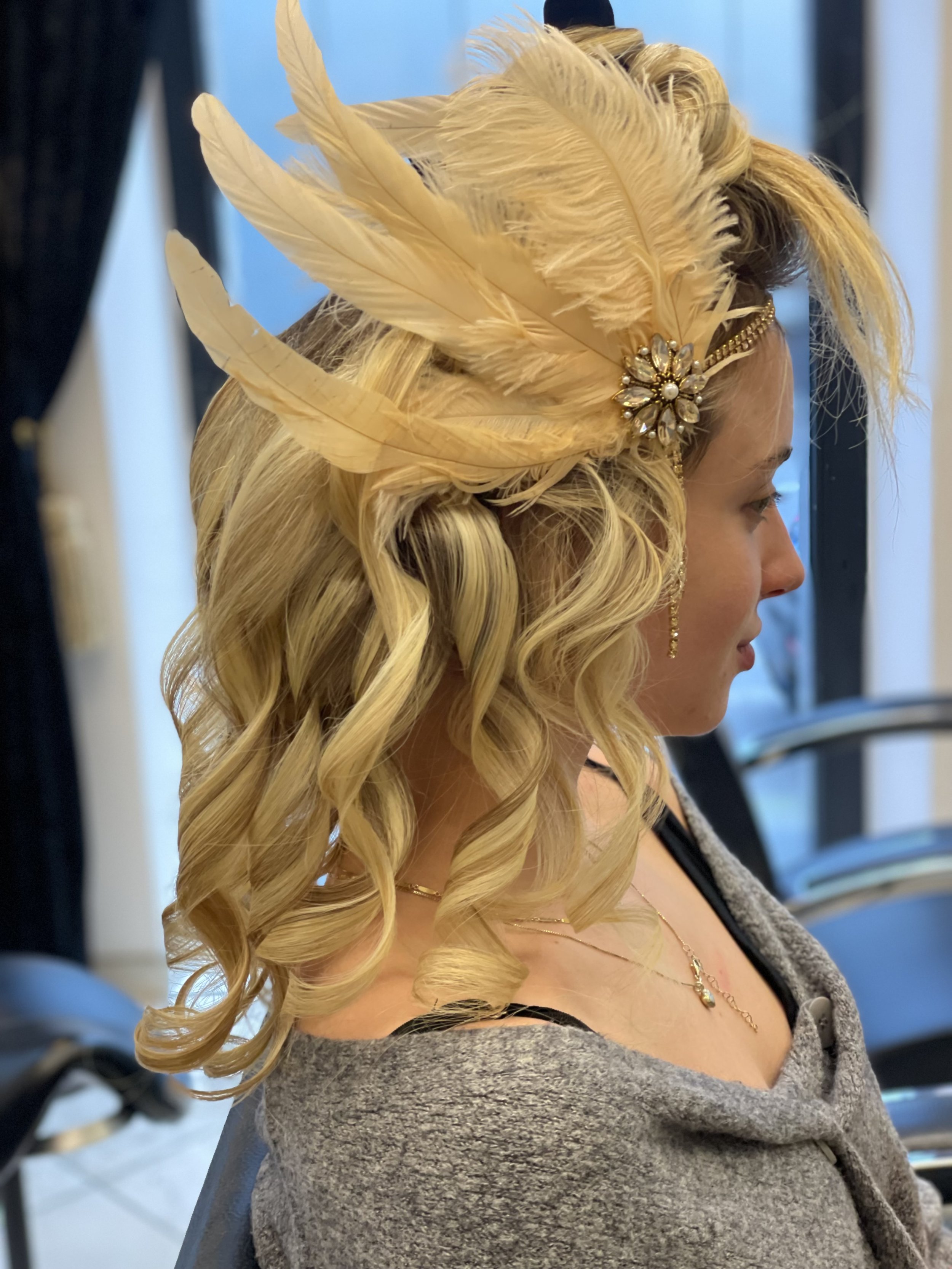 Frau mit eleganter Frisur und elegantem Haaraccessoire aus Federn und Schmuck, Schulteransicht, im Hintergrund dunkle und helle Vorhänge.