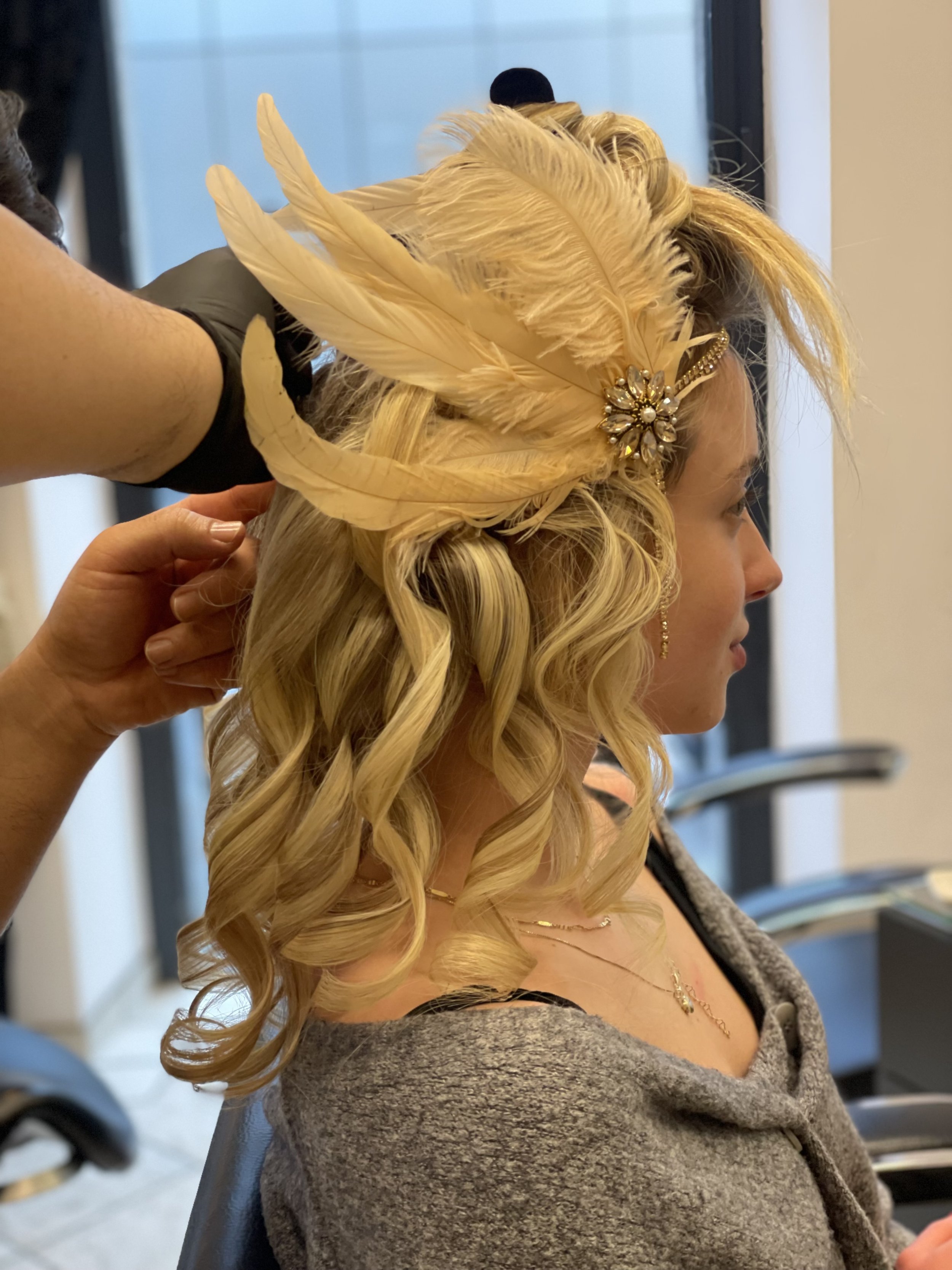 Frau mit blonden, lockigen Haaren und Federschmuck beim Friseurbesuch im Salon.