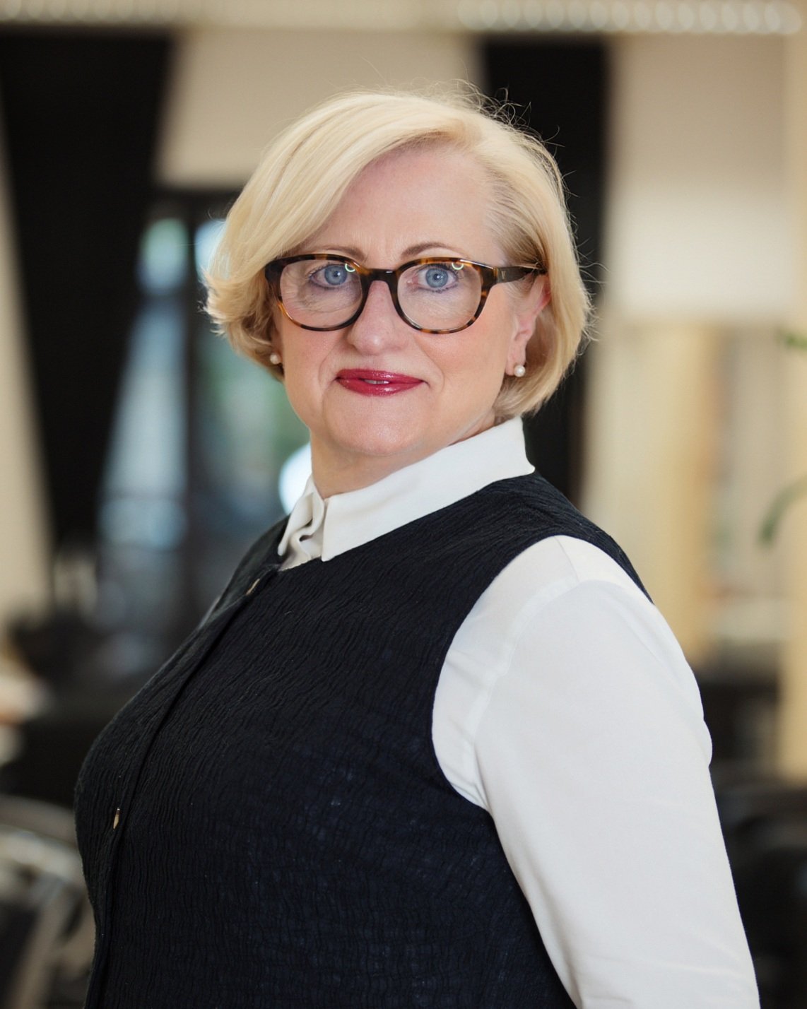 Porträt einer älteren Frau mit blondem Haar, Brille und rotem Lippenstift, trägt weiße Bluse und schwarze Weste, im Innenraum.