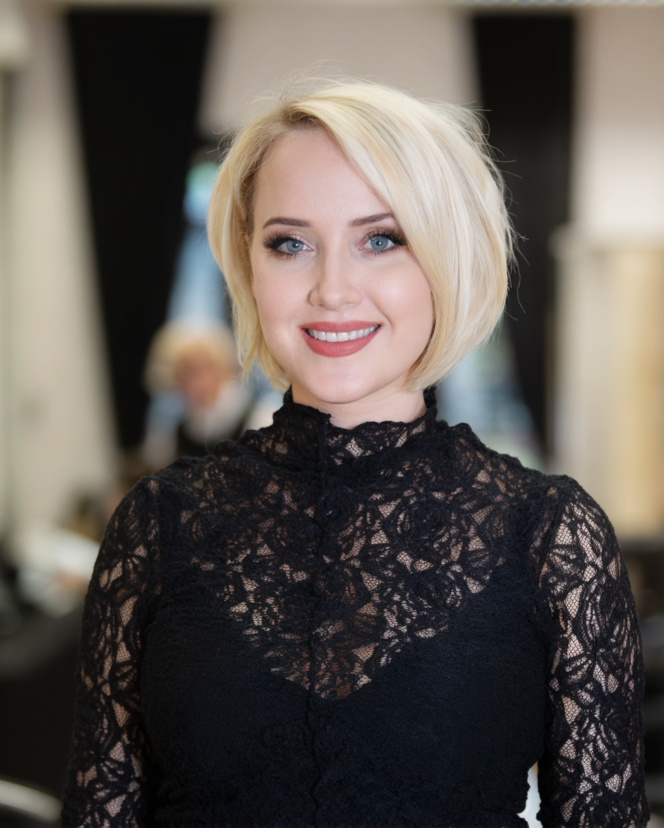 Junge Frau mit blondem, kurzem Haar trägt schwarze Spitzenbluse, lächelt vor unscharfem Hintergrund im Innenraum.