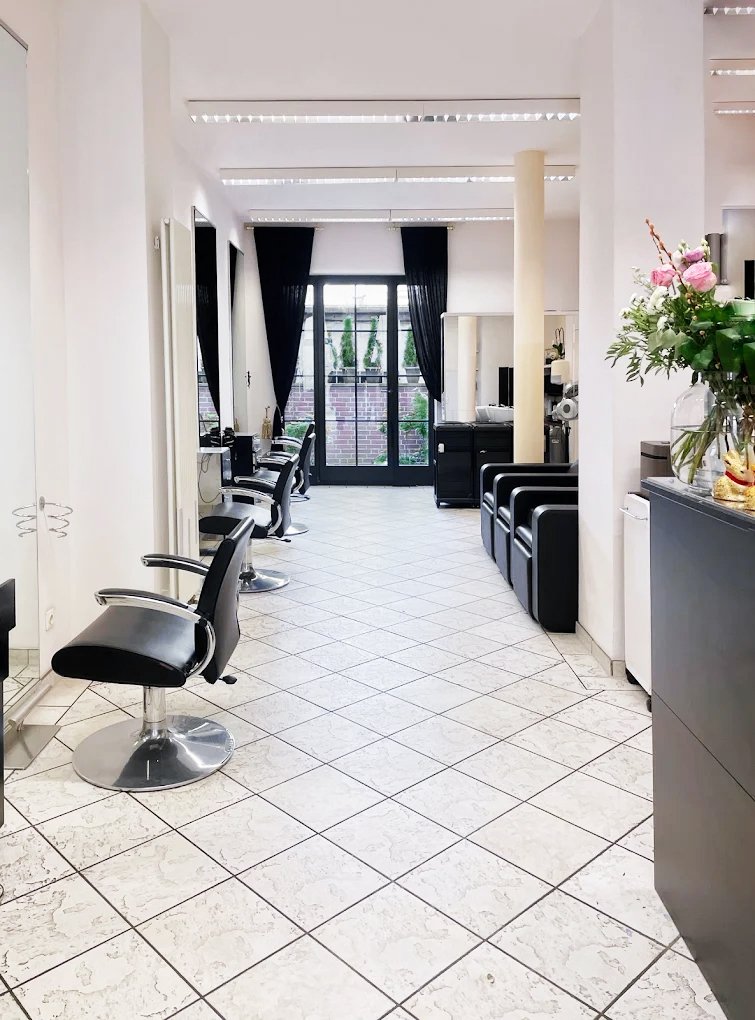 Friseursalon mit schwarzen Haarstühlen, Spiegeln und Waschstationen, große Fenster mit schwarzen Vorhängen, Fliesenboden, Blumen auf einem Regal.