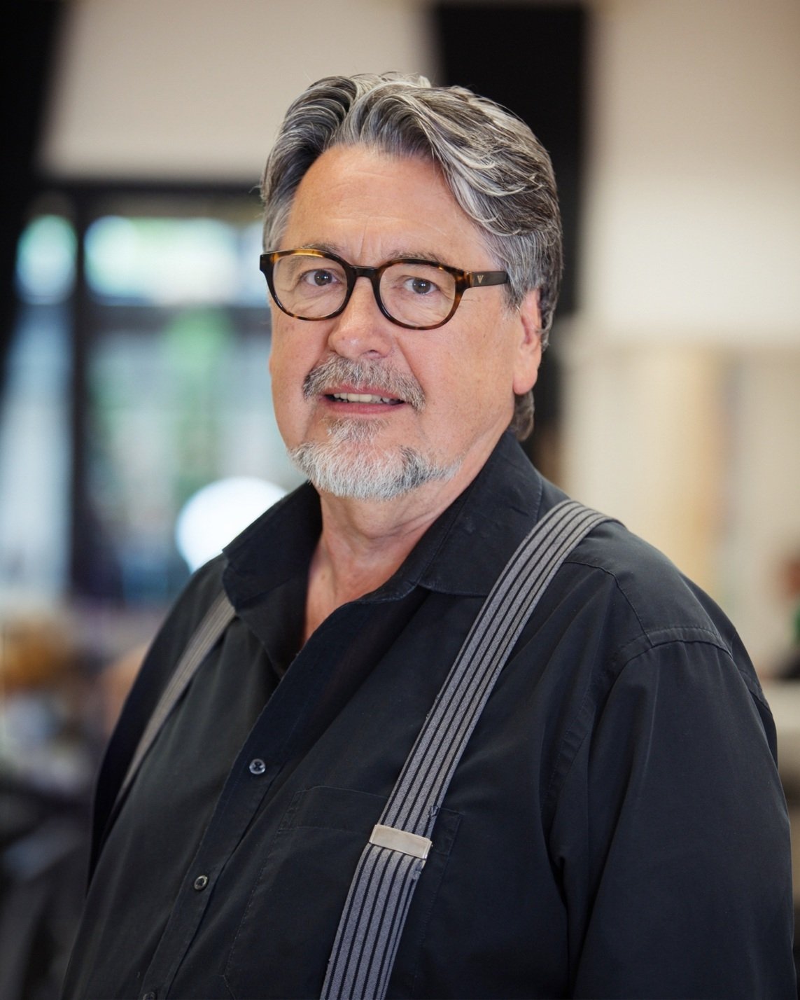Porträt eines älteren Mannes mit grauem Haar, Bart, Brille in schwarzem Hemd und Hosenträgern in einem Raum mit unscharfem Hintergrund.