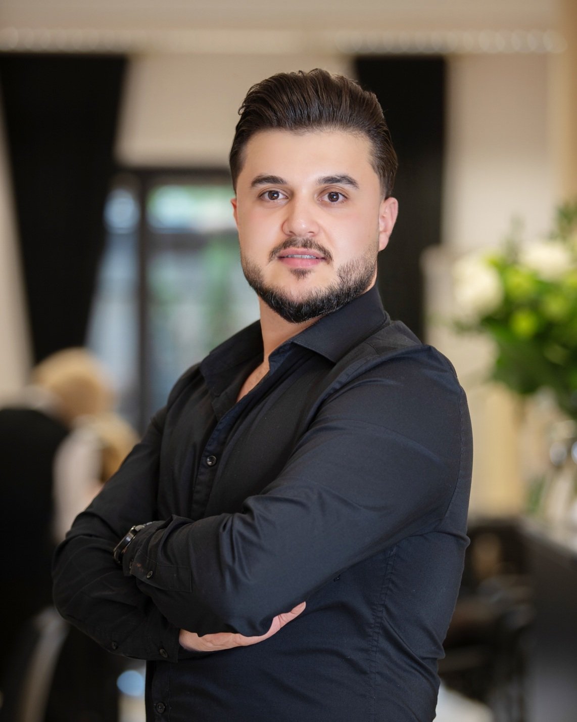 Ein junger Mann mit dunklem Haar und Bart, trägt schwarze Kleidung, steht mit verschränkten Armen in einem modernen, hell erleuchteten Raum.