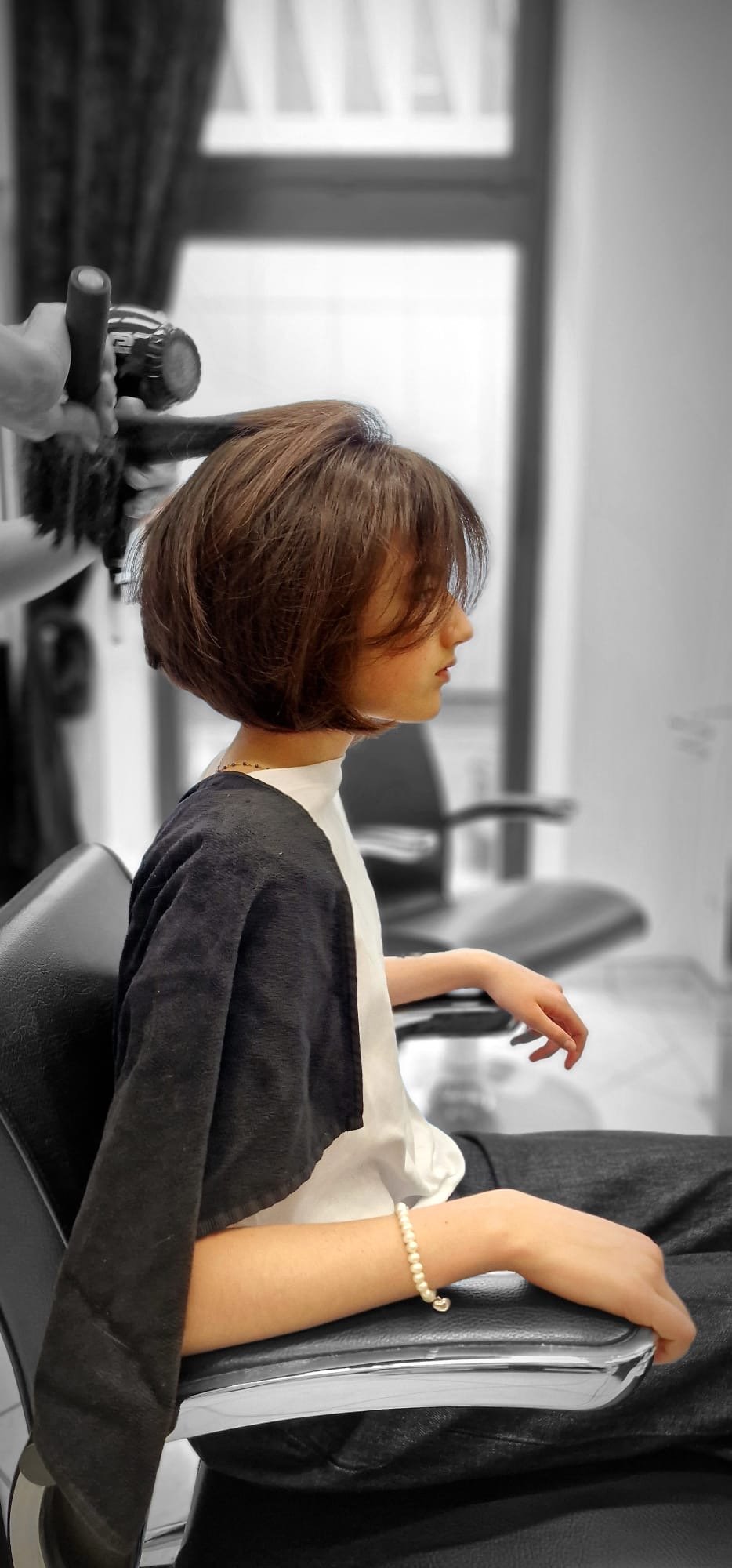 Frau bekommt im Friseursalon einen Haarschnitt, während sie auf einem schwarzen Friseurstuhl sitzt, mit braunen Haaren und einer Perlenarmband am Handgelenk.