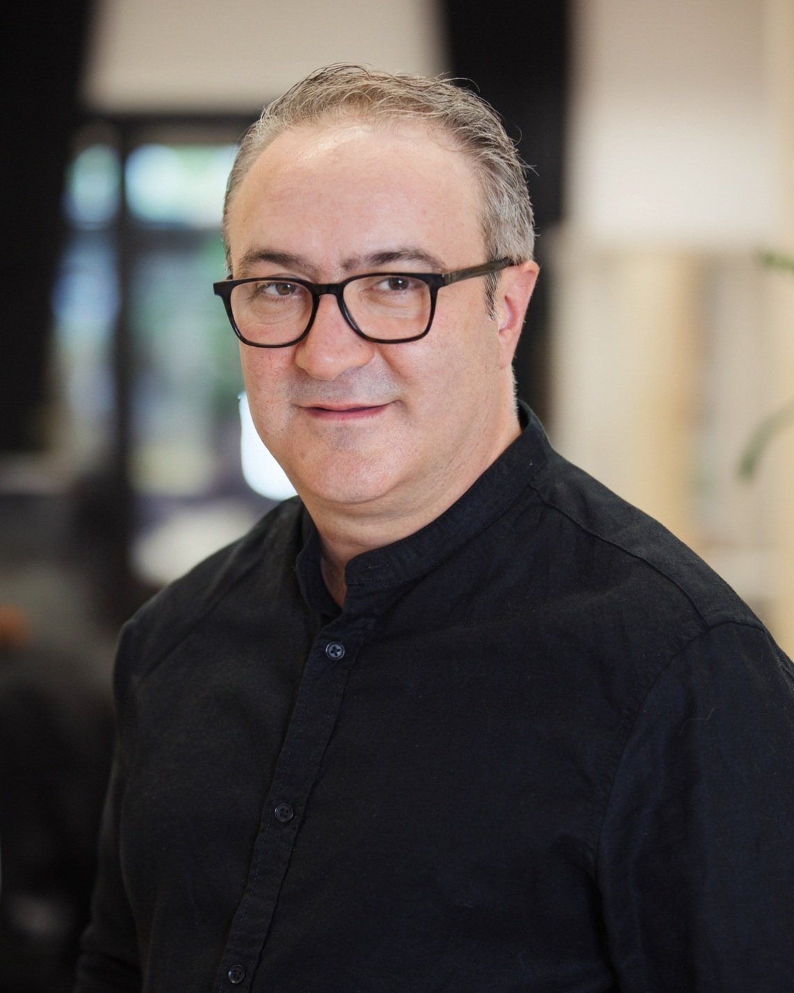 Porträt eines Mannes mit schwarzen Brillen und schwarzen Hemd, lächelnd, im Innenraum, unscharfer Hintergrund.